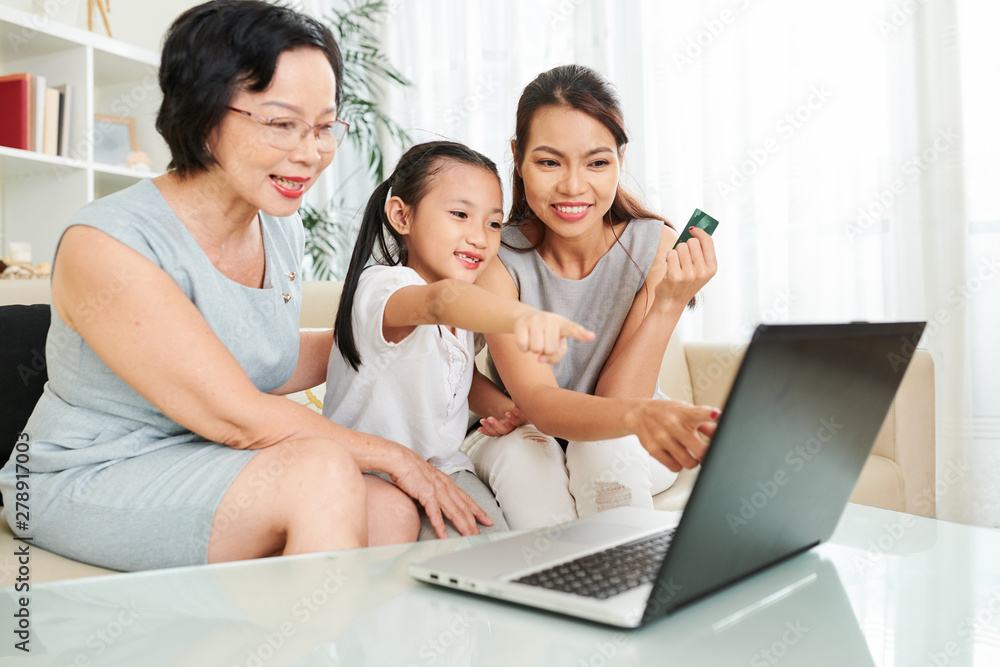 Happy Asian family sitting on sofa in front of the laptop computer pointing at it and they are going to do shopping online with credit card Happy Asian family sitting on sofa in front of the laptop computer pointing at it and they are going to do shopping online with credit card