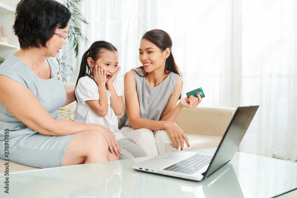 Asian little girl sitting together with mother and grandmother and looking at laptop with surprise they buying something with credit card online - obrazy, fototapety, plakaty 