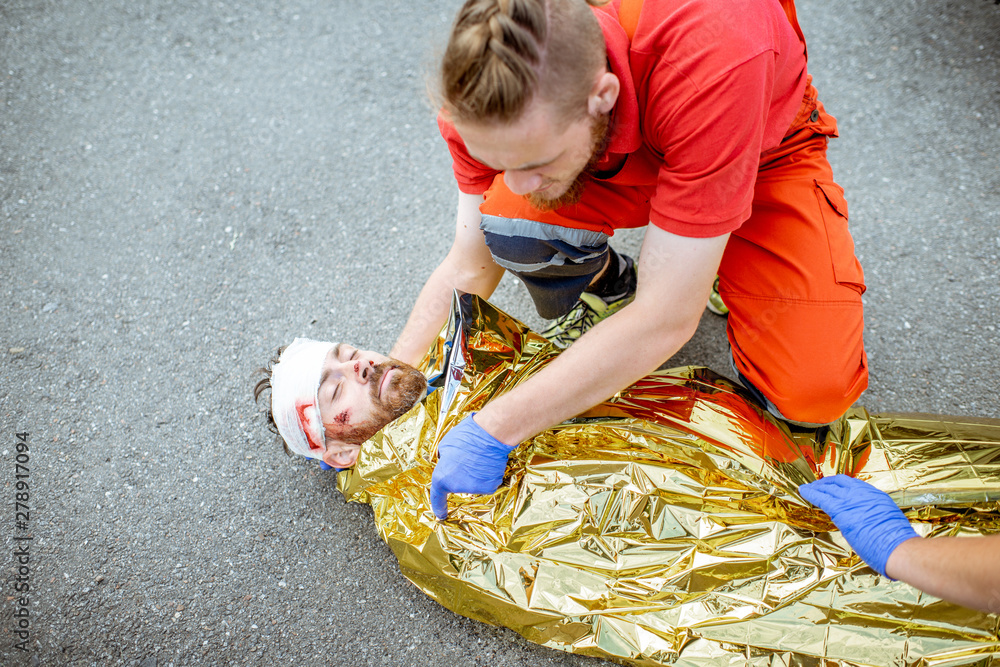 Fototapeta premium Ambluence worker covering injured man with thermal blanket, providing emergency care after the road accident