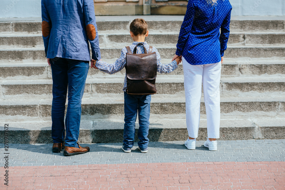 Parents and pupil of primary school stands hand in hand before stairs. Father, mother and son with backpack behind the back. Beginning of lessons. First day of fall. Back view