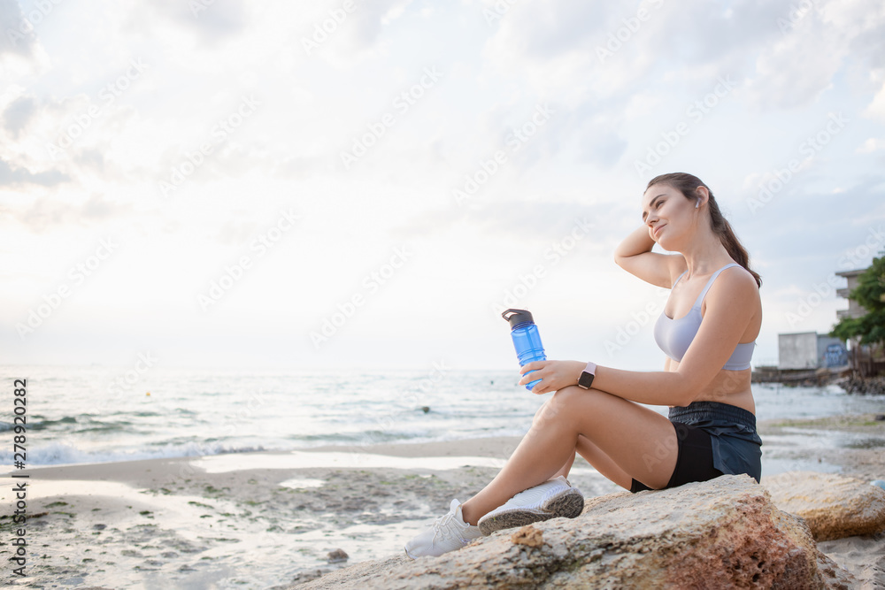 Young brunette woman with wireless earbuds and smart watches resting after morning workout at the sea shore at sunrise drinking water