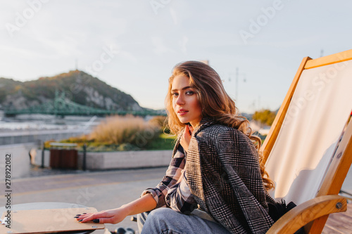 Sad girl with elegant hairstyle sitting in outdoor cafe on nature background. Attractive pensive woman with curly long hair waiting for her order in restaurant.