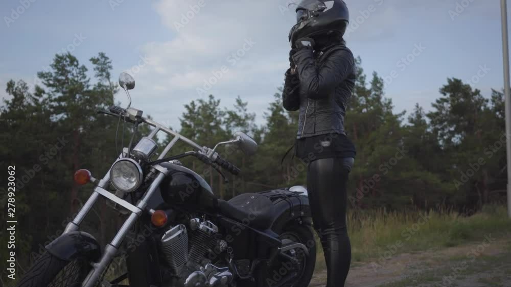 Skill caucasian woman in a black leather jacket and helmet riding a classic motorcycle. Girl removes the helmet standing near motorbike on the side of the road. Hobby, traveling and active lifestyle. Skill caucasian woman in a black leather jacket and helmet riding a classic motorcycle. Girl removes the helmet standing near motorbike on the side of the road. Hobby, traveling and active lifestyle.