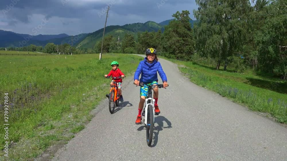 Two happy kids riding bikes on country road, beautiful nature with forest and mountains.