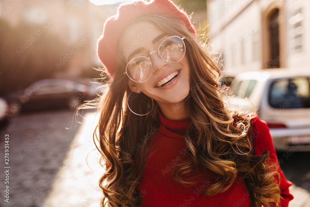 Close-up portrait of lovable french girl with elegant curly hairstyle looking to camera and laughing. Enthusiastic lovely lady in beret enjoying sunshine, walking on avenue. - obrazy, fototapety, plakaty 