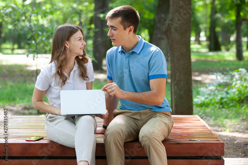 Young couple sitting together on bench in park and using laptop © biggur