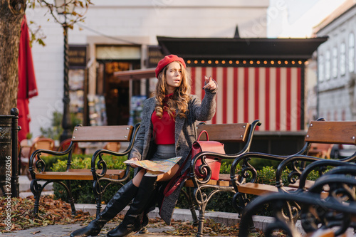 Serious curly woman looking away and pointing finger while sitting on bench with cafe on background. Outdoor photo of worried lady with city map in hands.