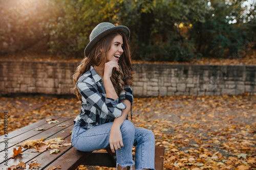 Romantic white woman with long brown hair looking away with dreamy smile, posing in sunny autumn day. Elegant curly female model with pale skin sitting on bench with green trees behind.