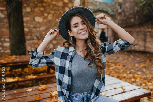 Fascinating european female model in casual checkered shirt posing with pleasure in yard in autumn. Dreamy white woman in hat enjoying september day.
