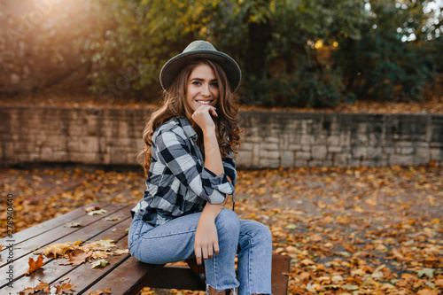 Spectacular brown-haired girl in casual clothes relaxing in park in good autumn weather. Refined white lady in gray elegant hat posing in sunny morning in yard, covered with fallen leaves.