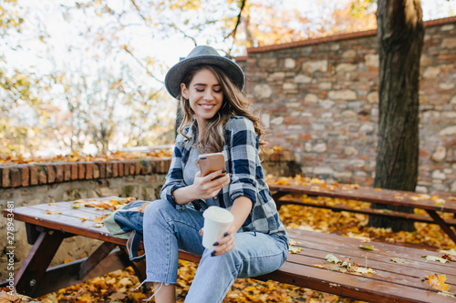 Magnificent white woman wears casual clothes texting message in good september day. Outdoor portrait of pretty girl with sincere smile using phone while chilling in autumn yard.