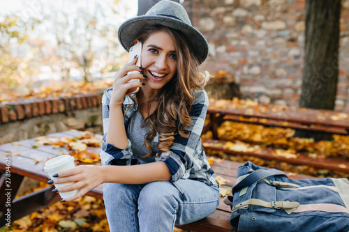 Stunning blue-eyed lady with curly hairstyle calling friend in good autumn day. Cute european girl in casual jeans talking on phone on autumn background.
