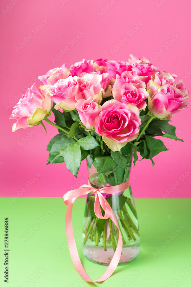 Bouquet of pink roses in a glass vase on a green and pink background. On a vase the red tape is tied. Bouquet of pink roses in a glass vase on a green and pink background. On a vase the red tape is tied.