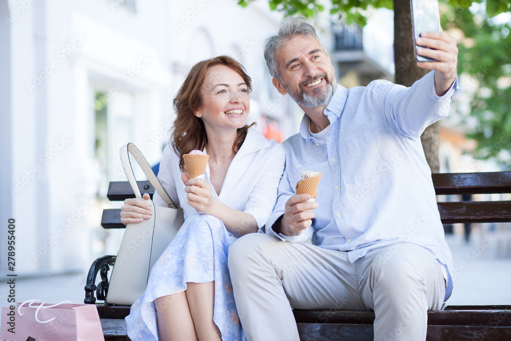 Smiling happy mature married couple sitting on a bench downtown, eating ice cream and posing for a selfie