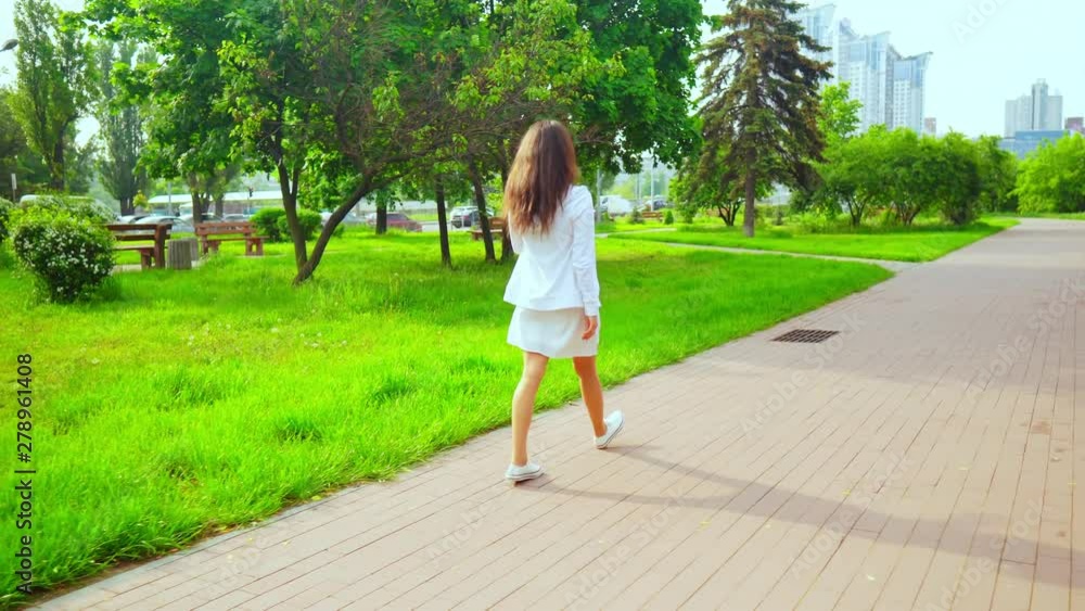 back view brunette wearing white dress and hoodie walking in modern town with green nature casual female going by the green trees on the background heavy traffic and city scape back view brunette wearing white dress and hoodie walking in modern town with green nature casual female going by the green trees on the background heavy traffic and city scape