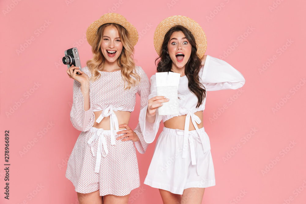 Happy friends women tourists posing isolated over pink wall background holding passport with tickets and camera. Happy friends women tourists posing isolated over pink wall background holding passport with tickets and camera.