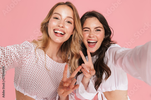 Smiling friends women posing isolated over pink wall background take selfie by camera.