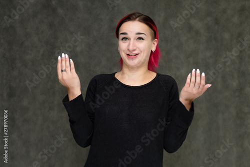 Portrait to the waist of a pretty girl with red hair on a gray background in a black jacket. Standing right in front of the camera in a studio with emotions, talking, showing hands, smiling