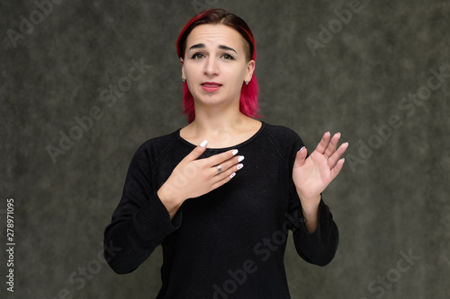 Portrait to the waist of a pretty girl with red hair on a gray background in a black jacket. Standing right in front of the camera in a studio with emotions, talking, showing hands, smiling