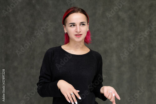 Portrait to the waist of a pretty girl with red hair on a gray background in a black jacket. Standing right in front of the camera in a studio with emotions, talking, showing hands, smiling