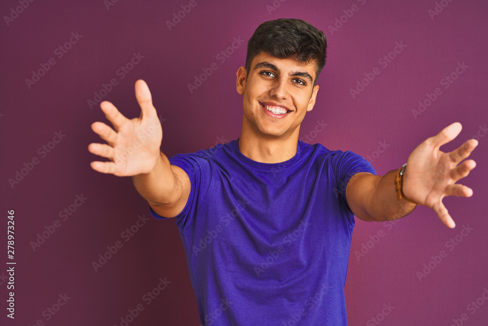 Young indian man wearing t-shirt standing over isolated purple background looking at the camera smiling with open arms for hug. Cheerful expression embracing happiness. Young indian man wearing t-shirt standing over isolated purple background looking at the camera smiling with open arms for hug. Cheerful expression embracing happiness.