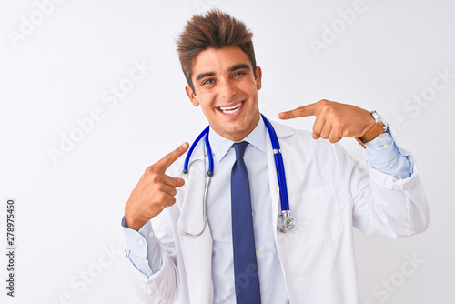 Young handsome doctor man wearing stethoscope over isolated white background smiling cheerful showing and pointing with fingers teeth and mouth. Dental health concept.
