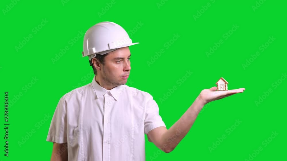 Young male smiling engineer in white hardhat and shirt standing and showing hand palm with small model of house on green screen background. Chroma key Young male smiling engineer in white hardhat and shirt standing and showing hand palm with small model of house on green screen background. Chroma key