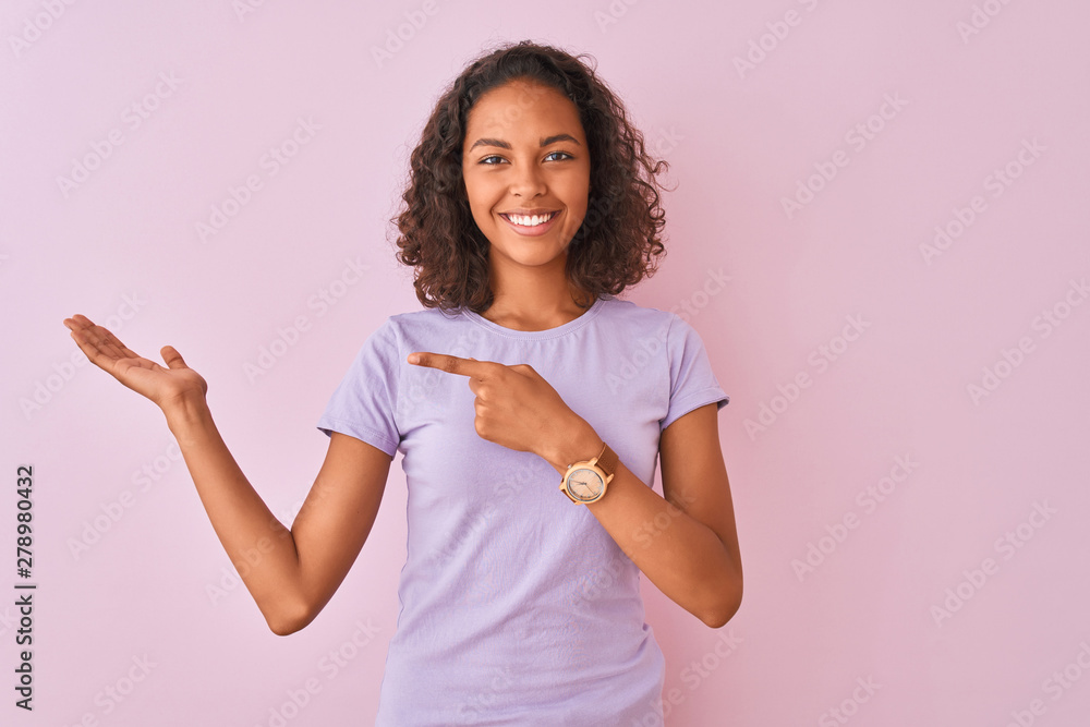 Young brazilian woman wearing t-shirt standing over isolated pink background amazed and smiling to the camera while presenting with hand and pointing with finger. Young brazilian woman wearing t-shirt standing over isolated pink background amazed and smiling to the camera while presenting with hand and pointing with finger.
