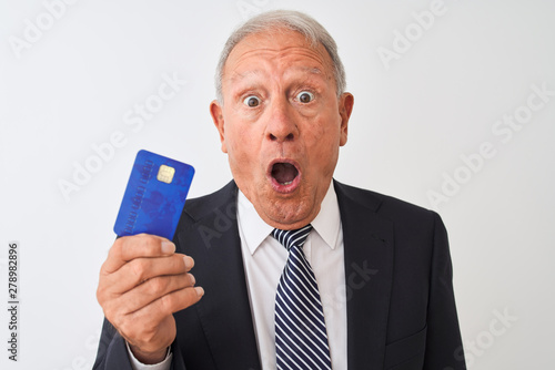 Senior grey-haired businessman holding credit card over isolated white background scared in shock with a surprise face, afraid and excited with fear expression © Krakenimages.com