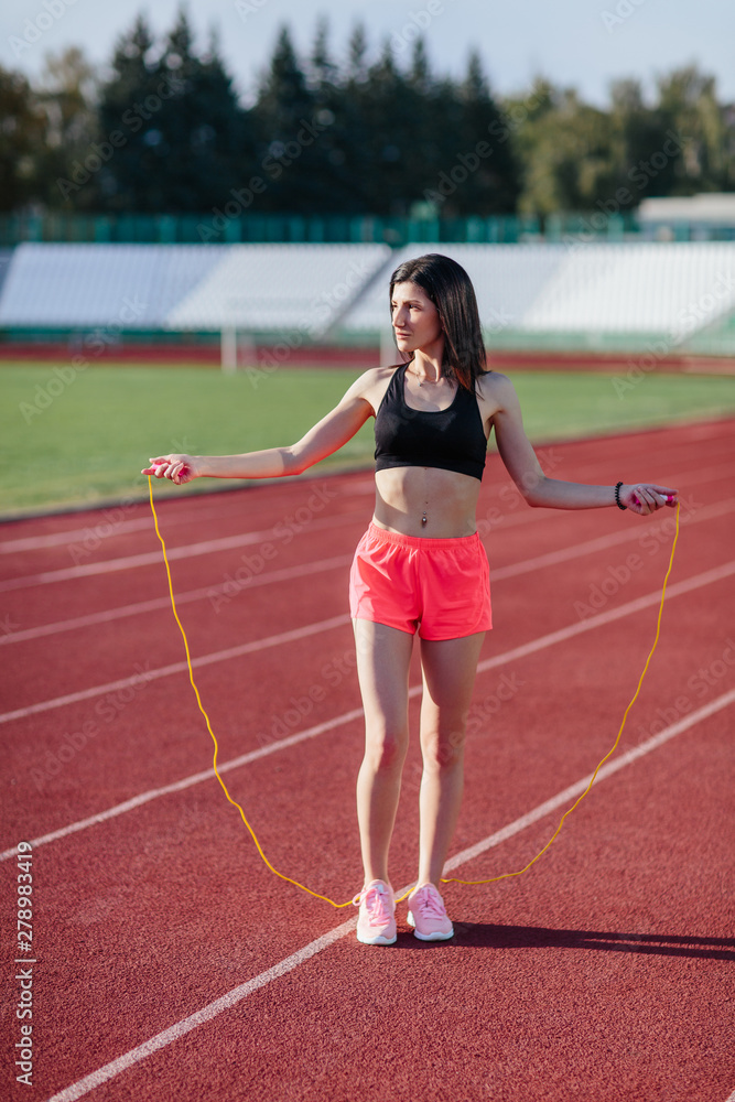 Young brunette woman athlete in pink shorts and black top on stadium sporty lifestyle standing on track holding jump rope under foot ready for workout