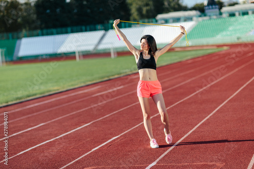 Portrait of happy young sports brunette woman in black top and rose shorts outdoors on stadium holding skipping rope over the head. © lena_itzy