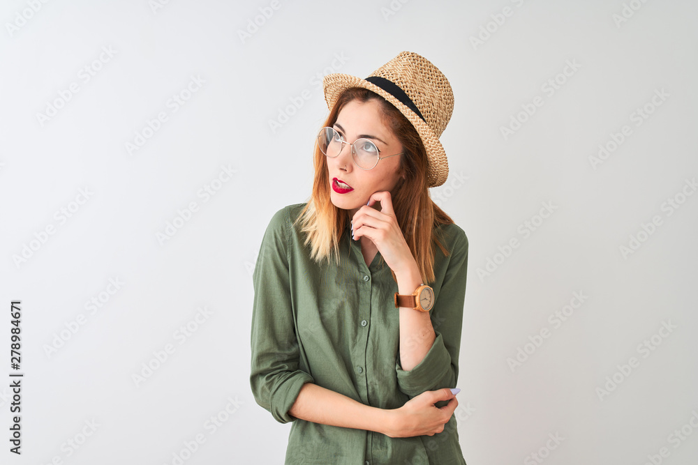 Redhead woman wearing green shirt glasses and hat over isolated white background with hand on chin thinking about question, pensive expression. Smiling with thoughtful face. Doubt concept. Redhead woman wearing green shirt glasses and hat over isolated white background with hand on chin thinking about question, pensive expression. Smiling with thoughtful face. Doubt concept.