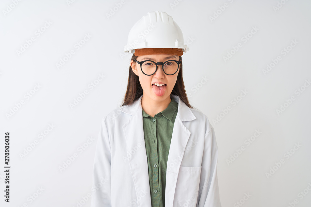 Young chinese engineer woman wearing coat helmet glasses over isolated white background sticking tongue out happy with funny expression. Emotion concept. Young chinese engineer woman wearing coat helmet glasses over isolated white background sticking tongue out happy with funny expression. Emotion concept.