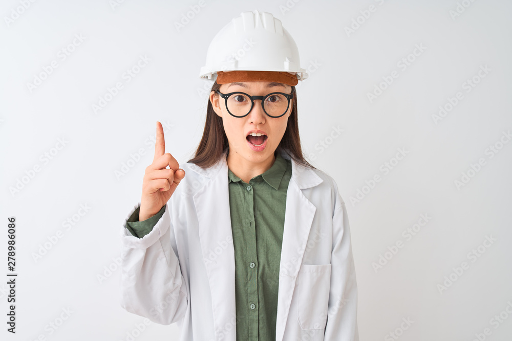 Young chinese engineer woman wearing coat helmet glasses over isolated white background pointing finger up with successful idea. Exited and happy. Number one. Young chinese engineer woman wearing coat helmet glasses over isolated white background pointing finger up with successful idea. Exited and happy. Number one.