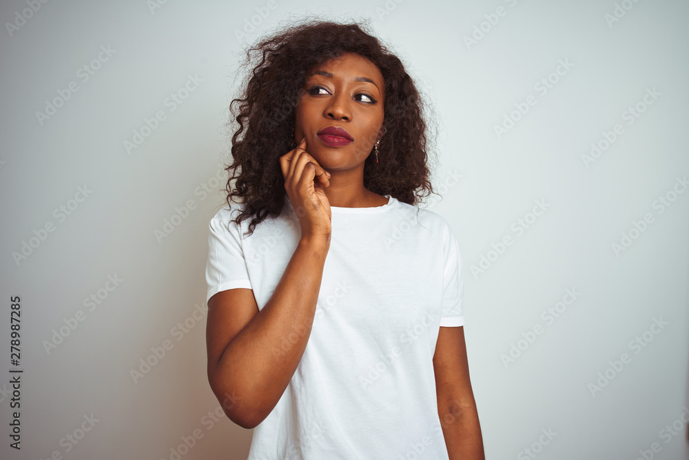 Young african american woman wearing t-shirt standing over isolated white background with hand on chin thinking about question, pensive expression. Smiling with thoughtful face. Doubt concept. Young african american woman wearing t-shirt standing over isolated white background with hand on chin thinking about question, pensive expression. Smiling with thoughtful face. Doubt concept.