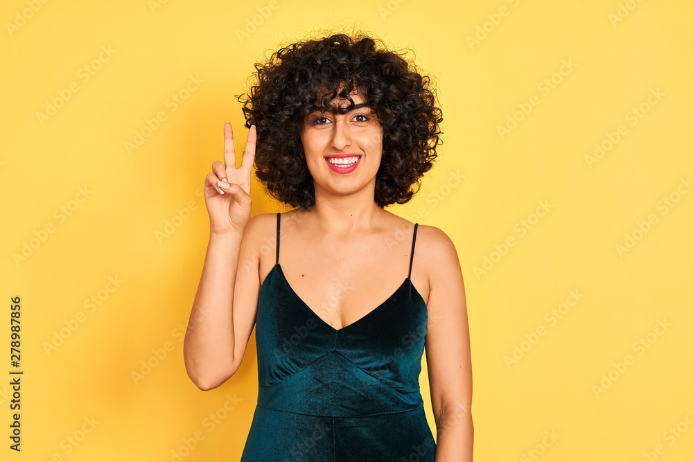 Young arab woman with curly hair wearing elegant dress over isolated yellow background showing and pointing up with fingers number two while smiling confident and happy. Young arab woman with curly hair wearing elegant dress over isolated yellow background showing and pointing up with fingers number two while smiling confident and happy.