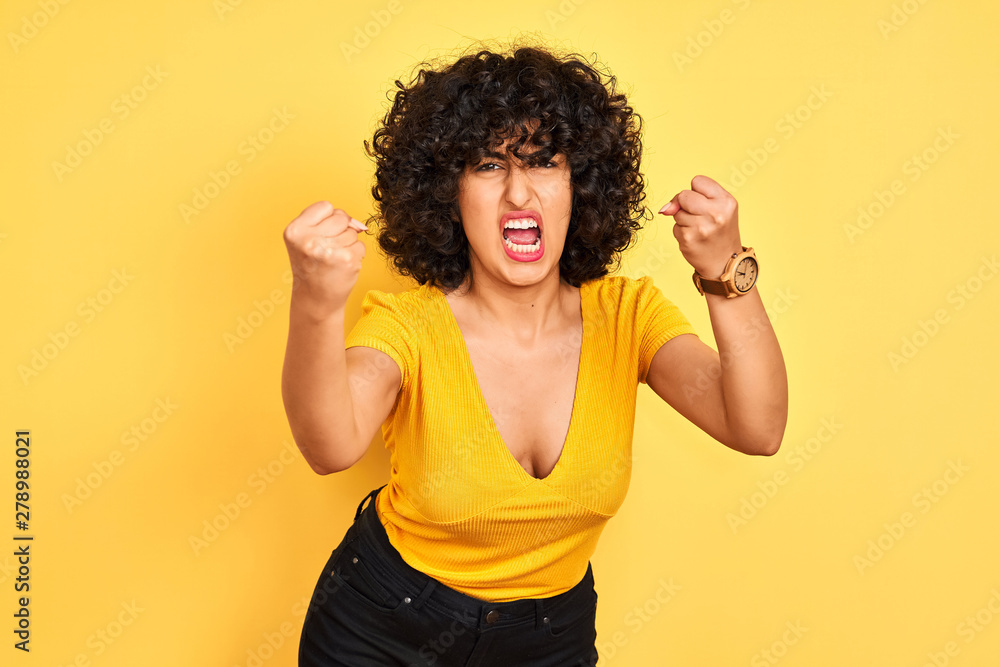 Young arab woman with curly hair wearing t-shirt standing over isolated yellow background angry and mad raising fists frustrated and furious while shouting with anger. Rage and aggressive concept. Young arab woman with curly hair wearing t-shirt standing over isolated yellow background angry and mad raising fists frustrated and furious while shouting with anger. Rage and aggressive concept.