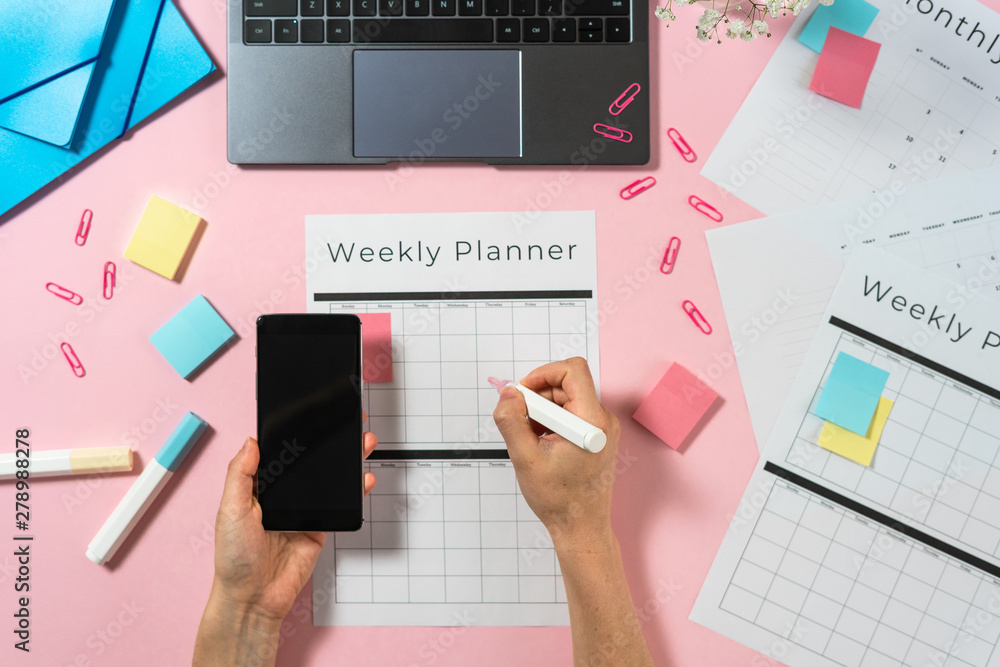 Woman holding smartphone and writing at planner on pink pastel background with laptop and stationery Woman holding smartphone and writing at planner on pink pastel background with laptop and stationery
