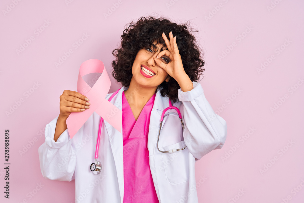 Young arab doctor woman with curly hair holding cancer ribbon over isolated pink background with happy face smiling doing ok sign with hand on eye looking through fingers Young arab doctor woman with curly hair holding cancer ribbon over isolated pink background with happy face smiling doing ok sign with hand on eye looking through fingers