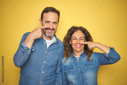 Beautiful middle age couple together standing over isolated yellow background Pointing with hand finger to face and nose, smiling cheerful. Beauty concept
