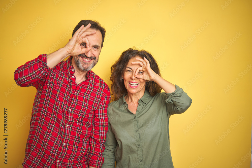 Beautiful middle age couple over isolated yellow background doing ok gesture with hand smiling, eye looking through fingers with happy face. Beautiful middle age couple over isolated yellow background doing ok gesture with hand smiling, eye looking through fingers with happy face.