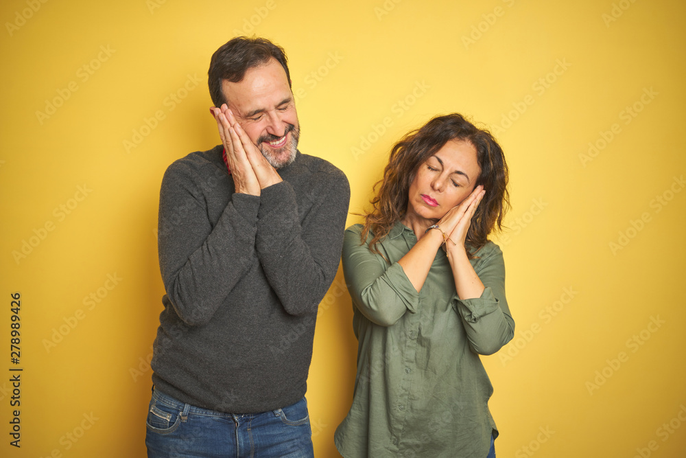 Beautiful middle age couple over isolated yellow background sleeping tired dreaming and posing with hands together while smiling with closed eyes. Beautiful middle age couple over isolated yellow background sleeping tired dreaming and posing with hands together while smiling with closed eyes.