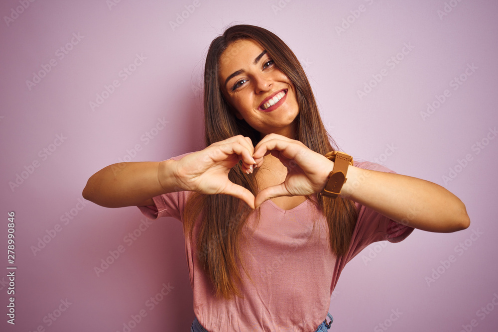 Young beautiful woman wearing t-shirt standing over isolated pink background smiling in love showing heart symbol and shape with hands. Romantic concept.