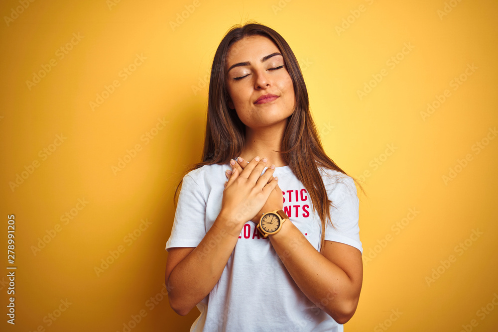 Obraz premium Beautiful woman wearing fanny t-shirt with irony comments over isolated yellow background smiling with hands on chest with closed eyes and grateful gesture on face. Health concept.