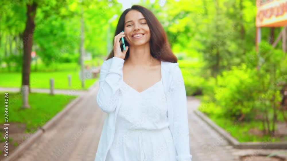 woman using smartphone chatting with friend walking on the street in park. Young female wearing white dress and hoodie stroll outdoors in summer season woman using smartphone chatting with friend walking on the street in park. Young female wearing white dress and hoodie stroll outdoors in summer season