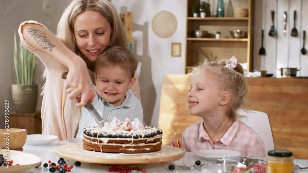 Waist-up shot of lovely young Caucasian mom sitting at kitchen table with two proudly smiling children, holding knife together with toddler son and slicing their fresh layered berry cake for eating Waist-up shot of lovely young Caucasian mom sitting at kitchen table with two proudly smiling children, holding knife together with toddler son and slicing their fresh layered berry cake for eating