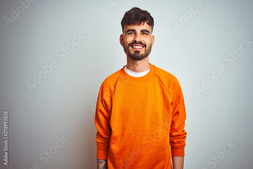 Obraz na plátně Young man with tattoo wearing orange sweater standing over isolated white background with a happy and cool smile on face