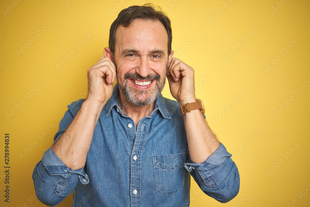 Handsome middle age senior man with grey hair over isolated yellow background covering ears with fingers with annoyed expression for the noise of loud music. Deaf concept. Handsome middle age senior man with grey hair over isolated yellow background covering ears with fingers with annoyed expression for the noise of loud music. Deaf concept.