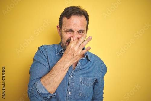 Handsome middle age senior man with grey hair over isolated yellow background smelling something stinky and disgusting, intolerable smell, holding breath with fingers on nose. Bad smells concept. © Krakenimages.com