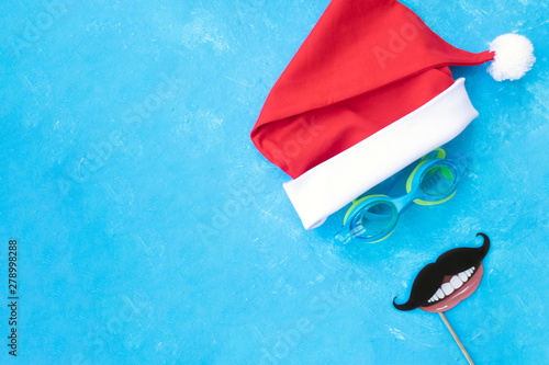 Hat of Santa Claus with goggles for swimming and the moustache and the smile. Christmas vacation, sandals and swimming glasses by water, slippers and pool goggles near swimming pool